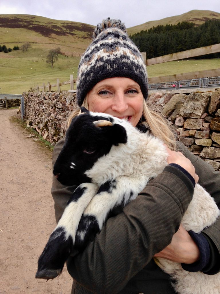 Angela Moffat, Stylist based in Edinburgh, Scotland, smiling while holding a black and white lamb during a farm visit in the Scottish Highlands