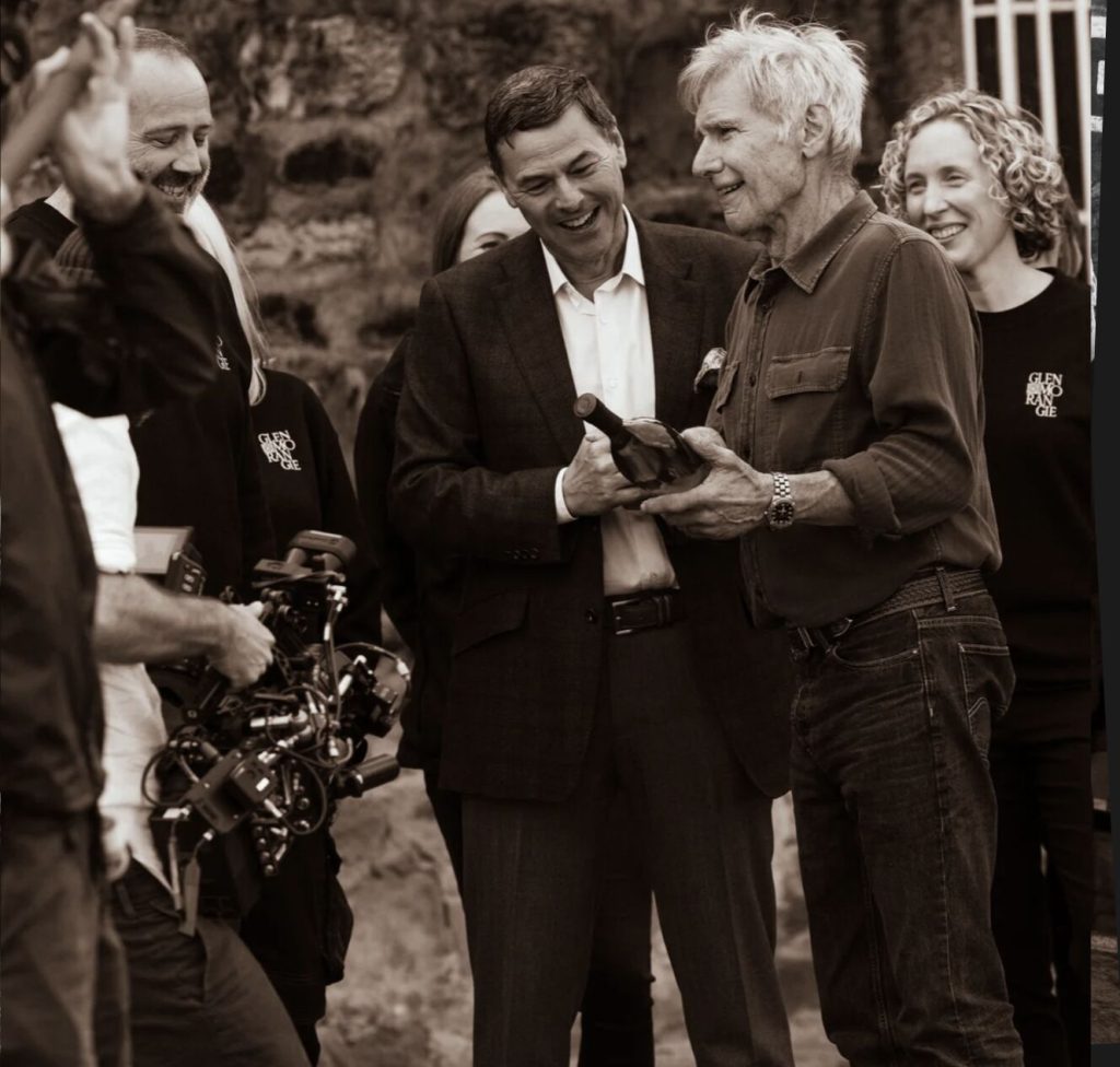 Harrison Ford receiving a bottle of Glenmorangie from Dr Bill Lumsden, of Distilling, Whisky Creation, and Whisky Stocks at Glenmorangie and Glenmorangie Distillery staff